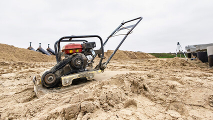 A compacting machine uses vibrations to compress sand on a construction site. Workers prepare the ground for building amidst cloudy skies in a rural area