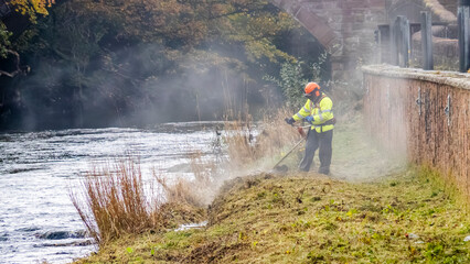 Obraz premium Environmental agency worker cutting grass on a riverbank to protect from flooding.