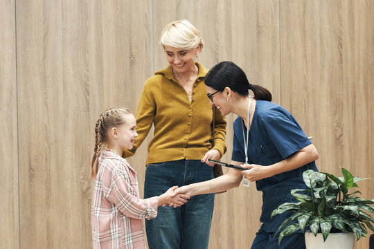 Caucasian girl shaking hands with Caucasian female healthcare worker while middle aged Caucasian woman standing nearby smiling, medical professional holding digital tablet