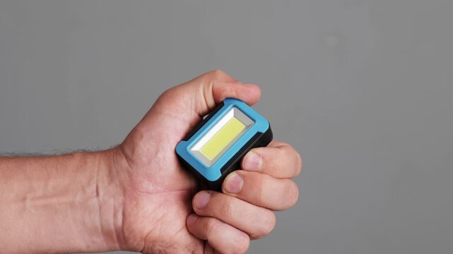A hand grips a small, portable LED work light, ideal for illuminating spaces during repairs or outdoor activities, showcasing its lightweight design
