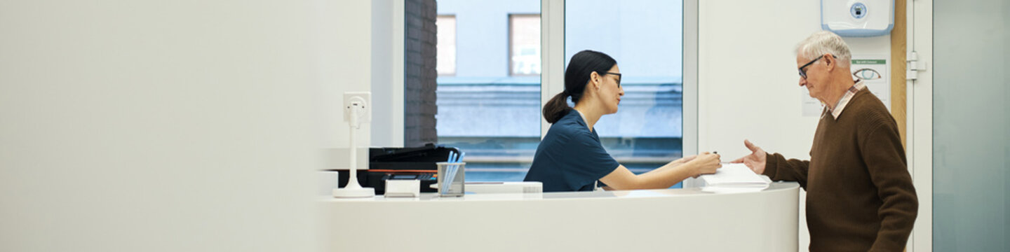 Header of Caucasian senior man interacting with young female nurse at reception desk in medical facility, woman handing document to man, both standing, modern healthcare environment visible