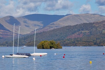 Fototapeta premium Lake scene with sailboats and an island. Mountains rise in the background, and buoys float in the calm water in Bowness-on-Windermere - Lake District - UK