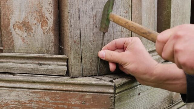 A person focuses on hammering a nail into a wooden corner joint, showcasing careful craftsmanship at a construction or repair site in bright daylight