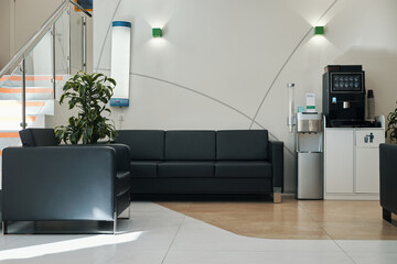 Empty modern waiting area showing black leather sofas, coffee machine, water dispenser and potted...