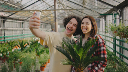 Young couple takes playful selfie while enjoying time together among lush plants in greenhouse setting during daylight