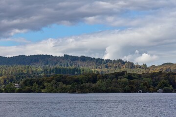 A landscape photo features a tranquil body of water in the foreground and a forested hillside under a cloudy sky in Bowness-on-Windermere - Lake District - UK