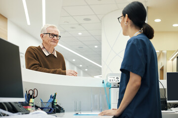 Obraz premium Senior Caucasian man standing at reception desk interacting with young adult Asian woman healthcare worker in medical facility, both engaged in conversation, modern clinic setting