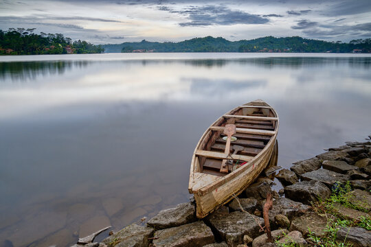 View of a weathered wooden boat resting on a rocky shore, mirroring the tranquil lake and distant hills under a soft, diffused sky, Brebes, Jawa Tengah, Indonesia.