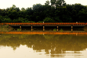 Long concrete bridge with orange safety railings crossing a calm muddy river with symmetrical reflections and dense forest background.