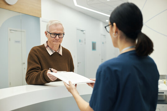 Senior Caucasian man standing at reception desk handing paperwork to young woman in medical scrubs, both interacting in modern healthcare facility interior