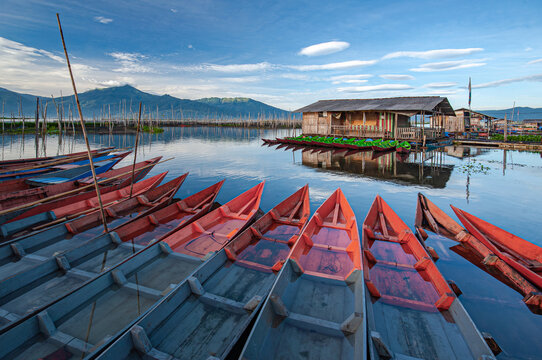 View of colorful boats lined up in the foreground leading to a wooden house on stilts over tranquil waters, Rawa Pening Lake, Ambarawa, Central Java, Indonesia.