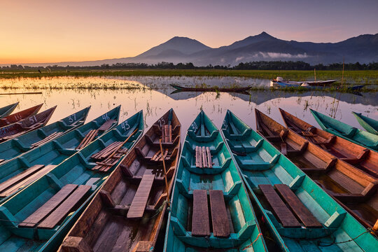 View of vibrant teal boats clustered together on the tranquil waters reflecting the mountain range at sunset, Ambarawa, Central Java, Indonesia.