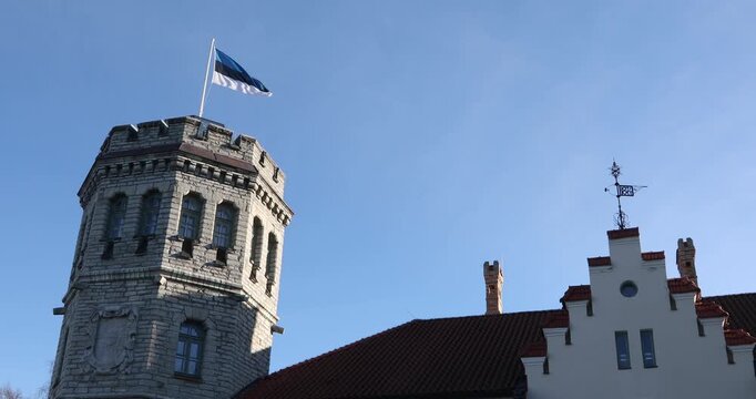 Close-up architectural view of a historic stone tower with the Estonian flag beside a stepped-gable roof and weather vane in Tallinn, Estonia. Crisp daylight, clear blue sky