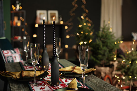 Holiday dinner table with black spiral candles, champagne flutes, gold napkins, and a redwhite runner, with small Christmas trees and string lights softly blurred in the background.