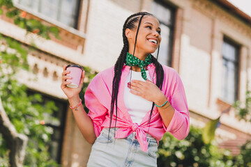 Happy young woman with braids wearing a pink jacket and green scarf holds a coffee cup smiles on a...