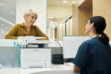 Caucasian middle aged woman smiling and talking to young adult woman receptionist at front desk in modern medical office, both women engaging in friendly conversation