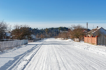 A wide, snow-covered road runs through a rural village in winter under a clear blue sky, flanked by...