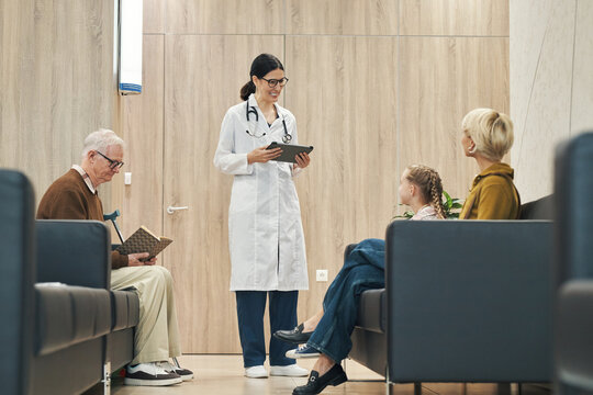 Middle aged Caucasian female doctor standing and holding digital tablet while talking to senior Caucasian man, middle aged Caucasian woman, and young Caucasian girl sitting in waiting room