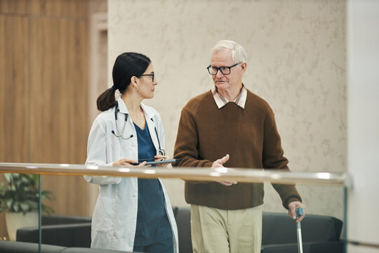 Middle aged Asian woman doctor walking and talking with senior Caucasian man holding cane in medical facility, both engaged in conversation, doctor holding digital tablet