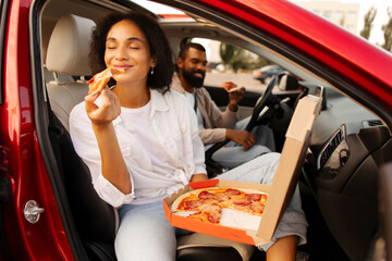 Satisfied Latin lady eating pizza sitting on passenger seat in car, holding cardboard box and...