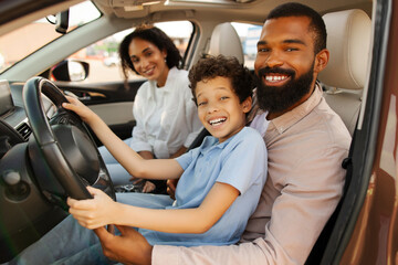 Father touching steering wheel with son sitting on his laps, and radiant mother looking at them...
