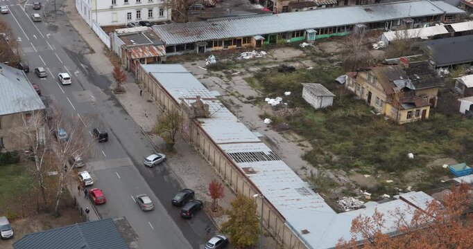 High-angle view of an abandoned industrial yard with rusted roofs, broken sheds and scattered trash amid puddles and weeds. Overcast light suggests neglect, hardship and slow transition.