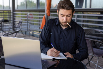 A man is sitting at a table with a laptop,holding a cigarette in his mouth.