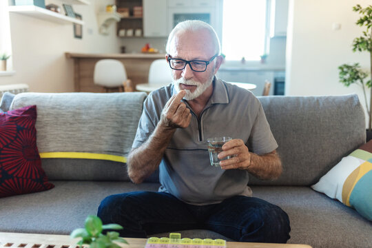 Smiling senior man sitting at a table in his home, taking daily medication from a blister pack while having a glass of water nearby, embracing a routine of health and wellness.
