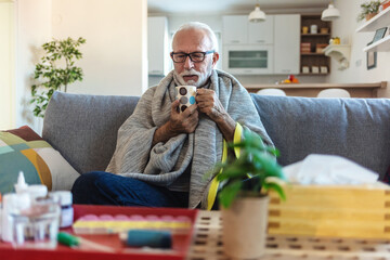 Senior man suffering from flu drinking tea while sitting wrapped in a blanket on the sofa at home....