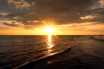 Spectacular Golden Sunset Over the Ocean Horizon with Dramatic Sunburst and Reflection on Dark Waves. Moody Seascape Photography Background Featuring a Solitary Silhouette Walking on the Wet Sand.