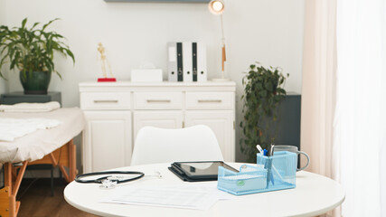 Bright Modern Medical Office Setup With Desk Tablet, Stethoscope, And Treatment Bed In Calm Clinic