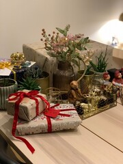 Cozy Christmas Gifts and Holiday Decorations on Table; wrapped gifts tied with red and gold ribbons, surrounded by festive decorations, pinecones, and houseplants on a wooden table