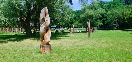 Abstract wooden sculptures stand tall in a lush green park, surrounded by trees and open grass,...