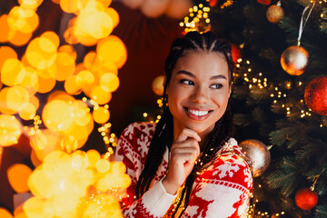 Whimsical christmas moment a smiling woman in a red white sweater beside a glowing tree and twinkling lights © deagreez