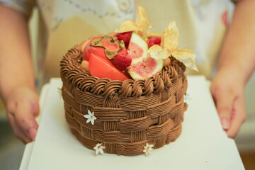 Two hands holding a chocolate cake box with a decorated cream cake topped with figs, persimmon,...