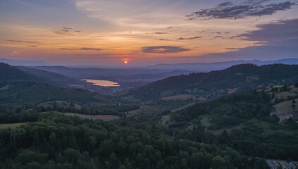 Bird's-eye perspective of a valley and mountain scene with water, summer vibes, natural surroundings, forested hills, sunset glow, farming fields, and ecological scenery