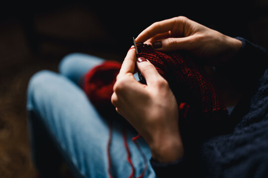 A person sits with jeans, knitting red yarn into a textured scarf. Close-up on hands guiding the needles conveys calm, focus, and handmade warmth perfect for craft, hobby, and gift ideas.