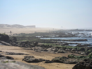 A beach scene with people walking on the sand and rocks in the foreground.