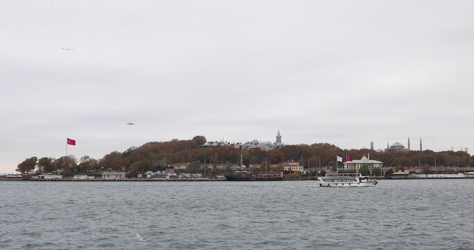Istanbul waterfront on an overcast day: tracking shot with a passenger ferry crosses the Bosphorus as Hagia Sophia and Topkapi hill rise behind, framed by autumn trees and Turkish flags. 