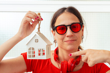 Confident woman wearing red shirt and sunglasses holding small white house model and looking at...