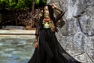 Mysterious woman in dramatic flowing black gown with gold mask posing by stone wall and tropical...