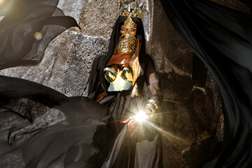 Mysterious woman in dramatic flowing black gown with gold mask posing by stone wall and tropical...