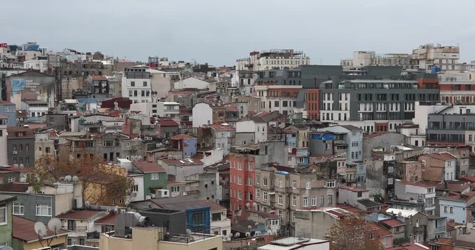 Wide view of a dense Istanbul neighborhood: layered apartment blocks, patched roofs, satellite dishes and air conditioners, with newer offices rising behind. Overcast light, history, everyday life.
