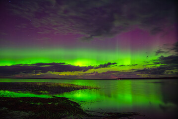 Vivid northern lights glowing green and purple over a calm lake with reflections and scattered clouds under a starry night sky.