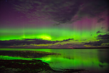 Vivid northern lights glowing green and purple over a calm lake with reflections and scattered clouds under a starry night sky.