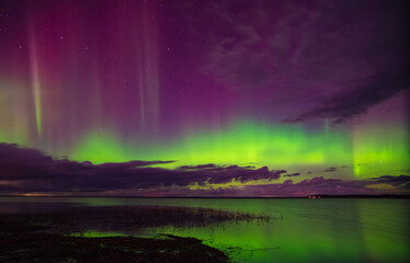 Brilliant aurora borealis in green and purple hues shines above a calm lake and cloudy night sky with faint stars.
