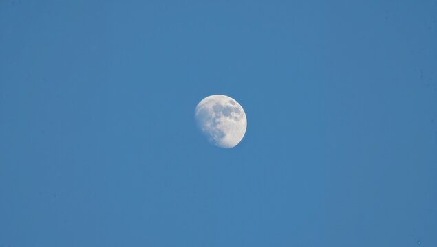 Wide shot of a large white moon against a bright blue daytime sky, showing nearly full illumination with a slight shadow from Earth. - Powered by Adobe