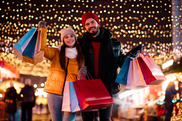 Funny festive couple enjoys shopping spree at outdoor market under twinkling lights during...