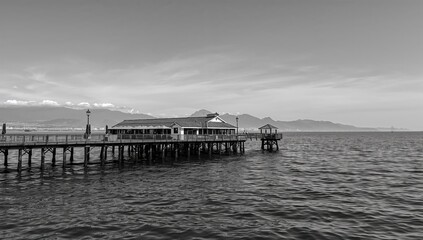 Obraz premium Coastal pier featuring dining establishments by the ocean shore. Monochrome setting with water, sky, beach, and summer vibes. Travel destination with isolated landscape and seaside restaurants.