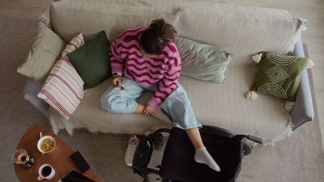 High angle shot of young woman in loungewear sitting on couch near wheelchair in modern living room and giving herself leg massage using triangle ball massager, recovering from illness
